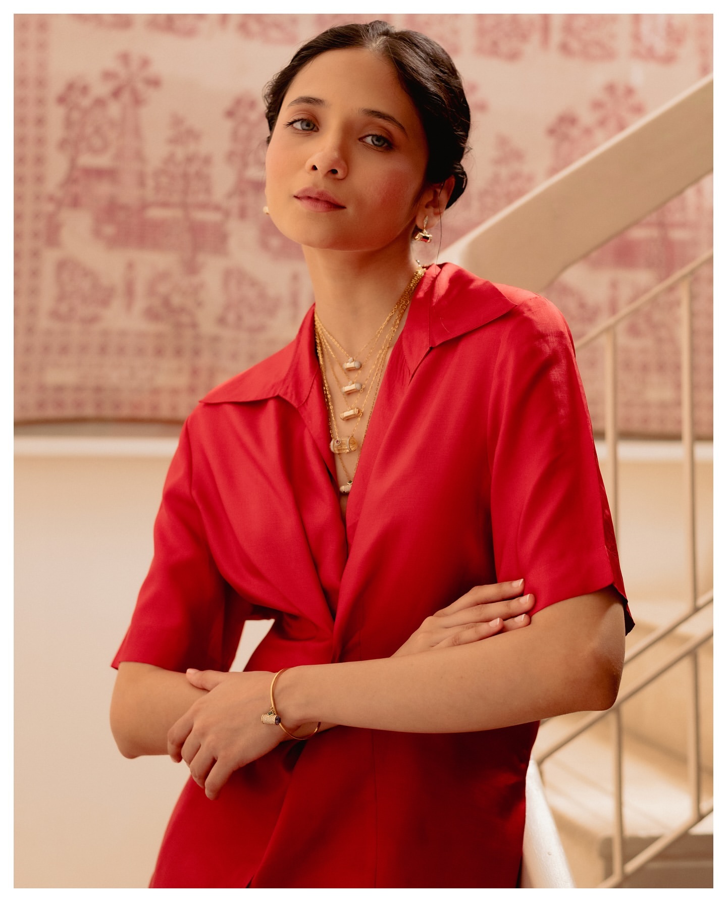 Portrait of a woman wearing a red shirt and gold jewelry, standing indoors with arms crossed, with a patterned background.