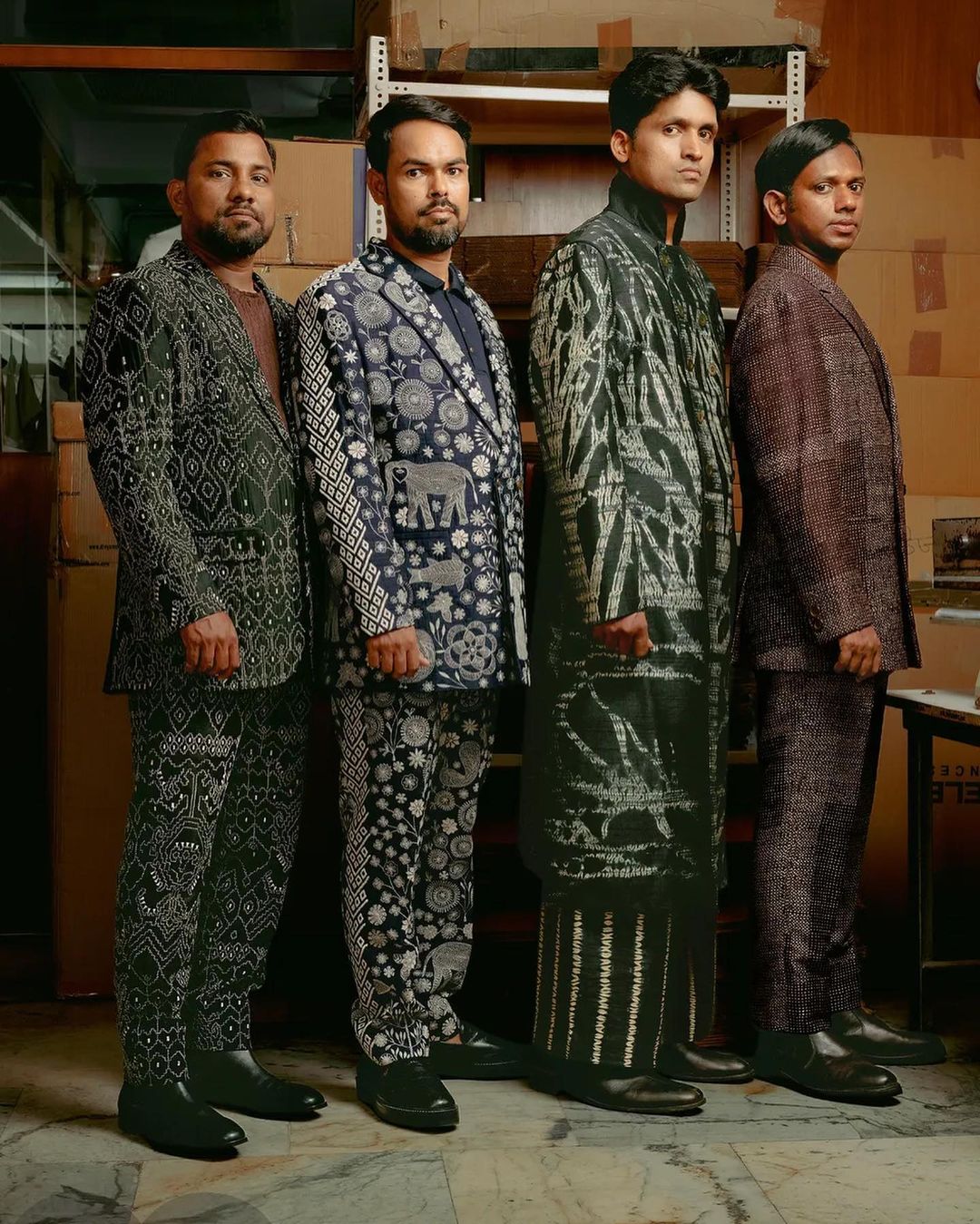 Four adult males standing in a row wearing patterned suits in an indoor environment featuring cardboard boxes in the background.