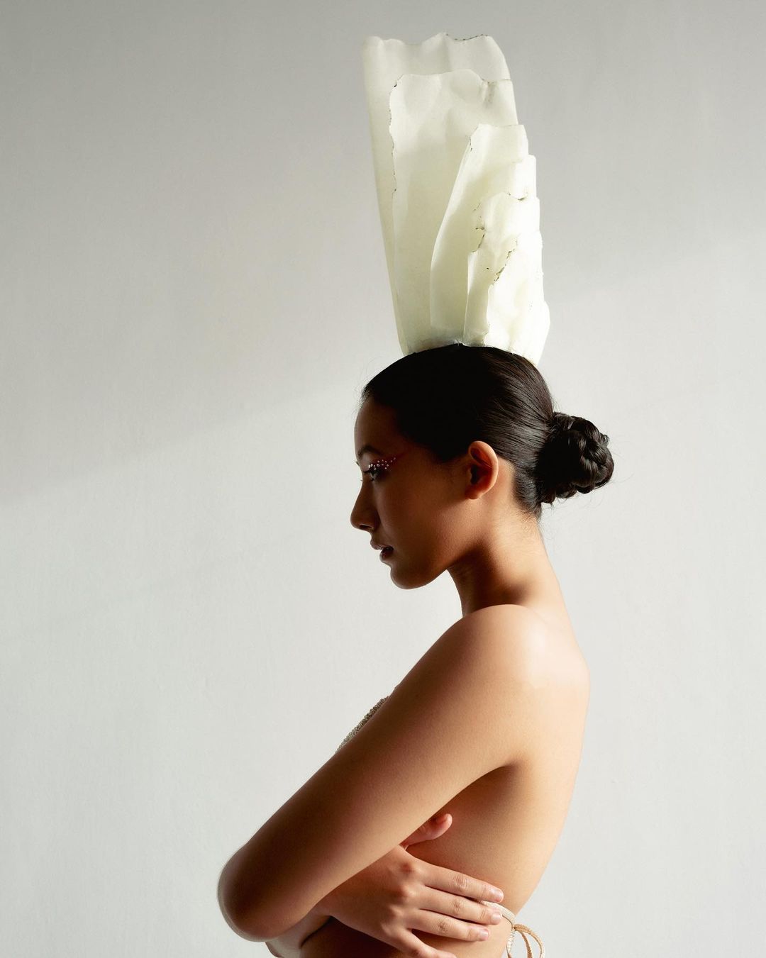Profile portrait of a woman with a stylized white headpiece, hands folded, soft lighting, and a minimalist white studio background.