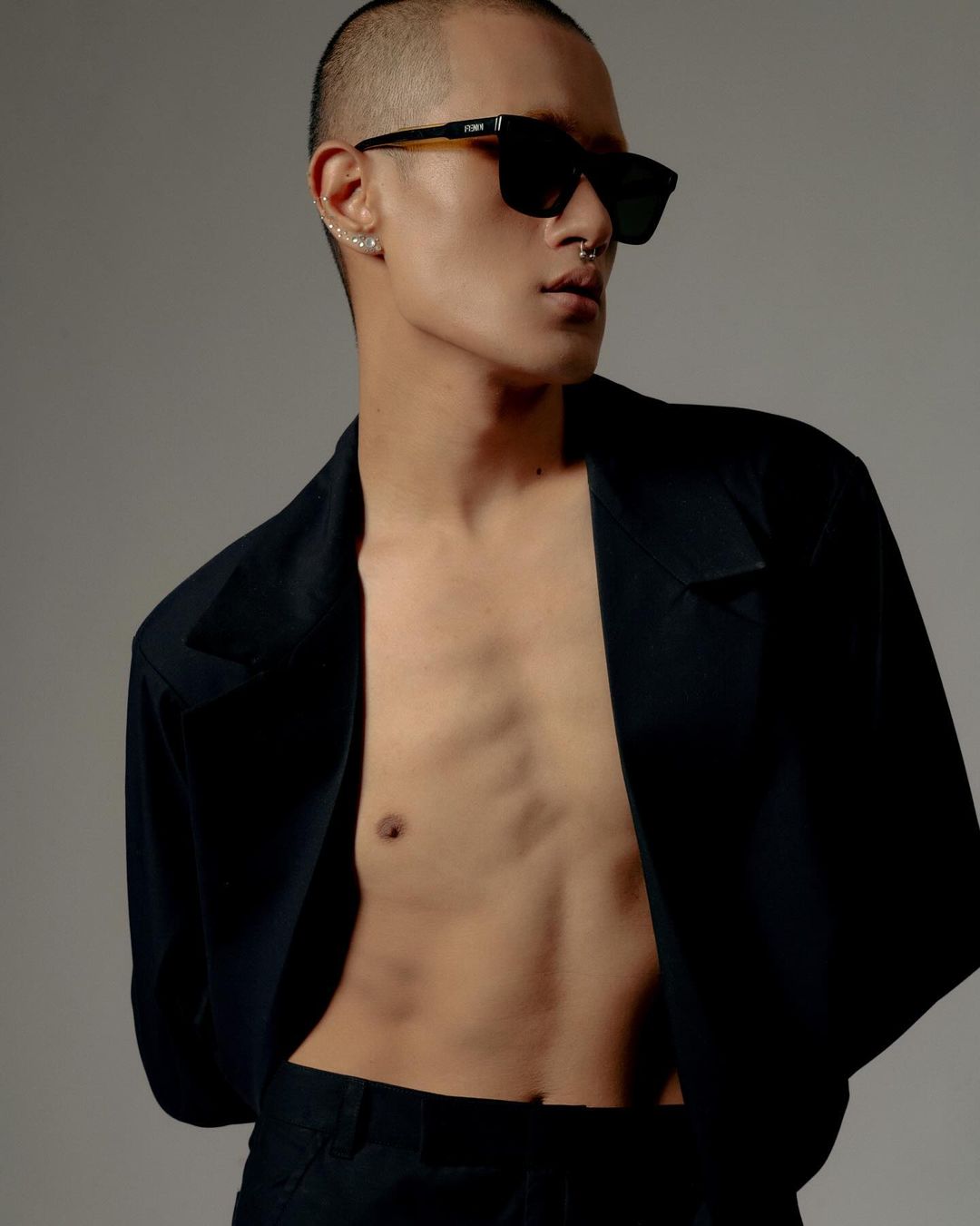 A shirtless adult male poses indoors wearing black sunglasses and a blazer, set against a neutral grey background.