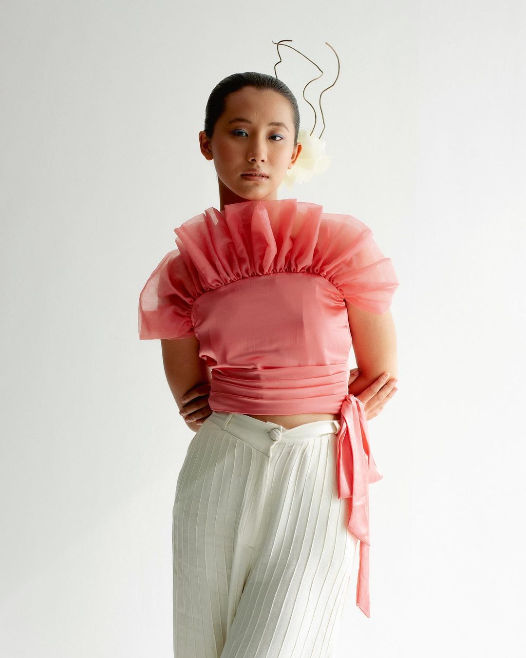 A portrait of an Asian woman in a pink blouse and white pants, posing in a light studio setting with a delicate floral headpiece.