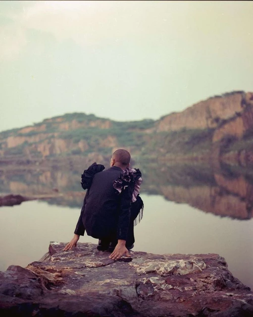 Person in black suit crouches on rock with water and mountains in background, facing away. Fashion editorial shot in natural light.