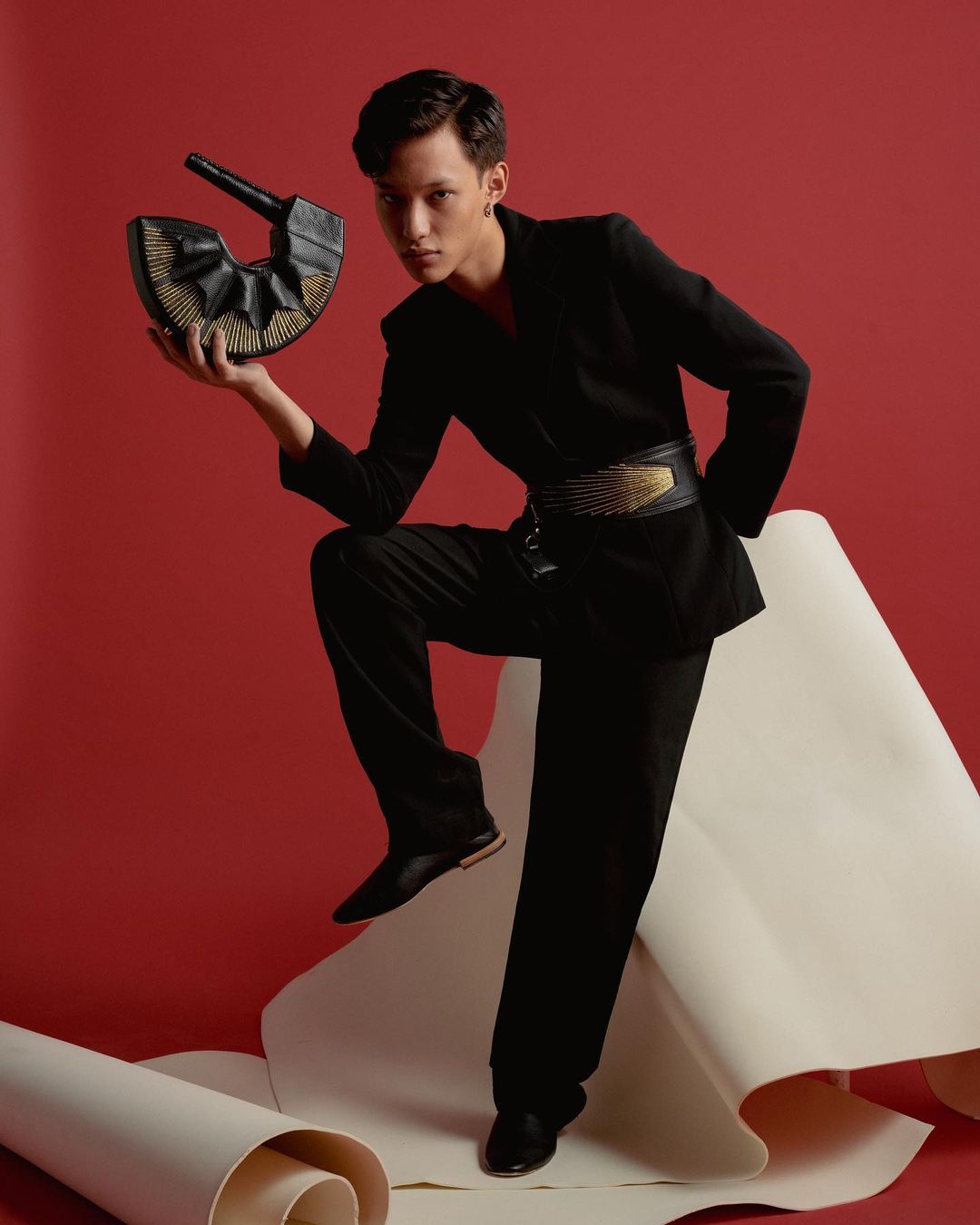 Adult male in a black suit poses in a studio with a red background, showcasing a black sculptural handbag and fashionable waist belt.
