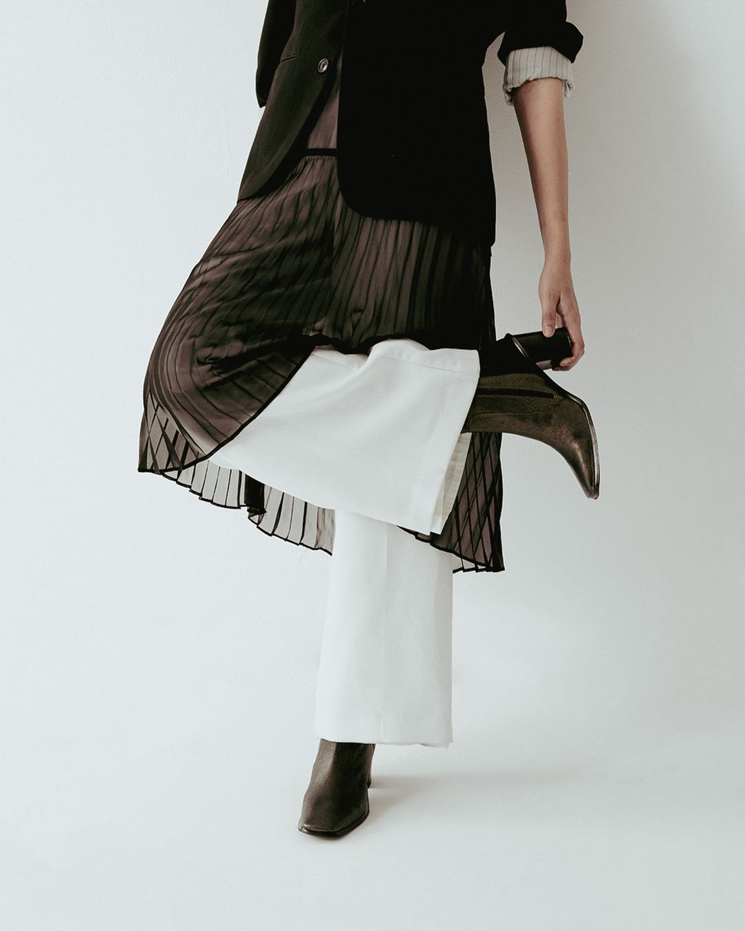 Fashionable woman in a black jacket, brown pleated skirt, white wide-leg pants, and brown heeled boots posing in a light studio.