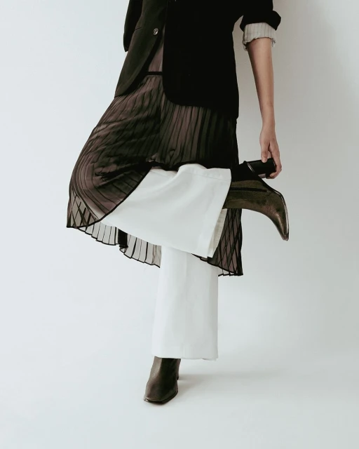 Fashionable woman in a black jacket, brown pleated skirt, white wide-leg pants, and brown heeled boots posing in a light studio.