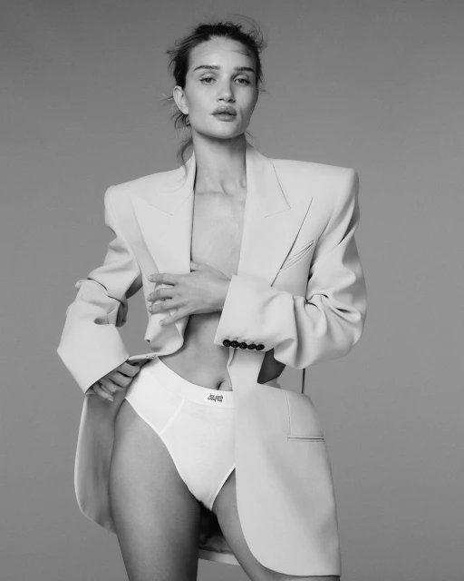 Black and white portrait of a confident woman in a blazer and underwear, posing in a photo studio with a neutral backdrop.