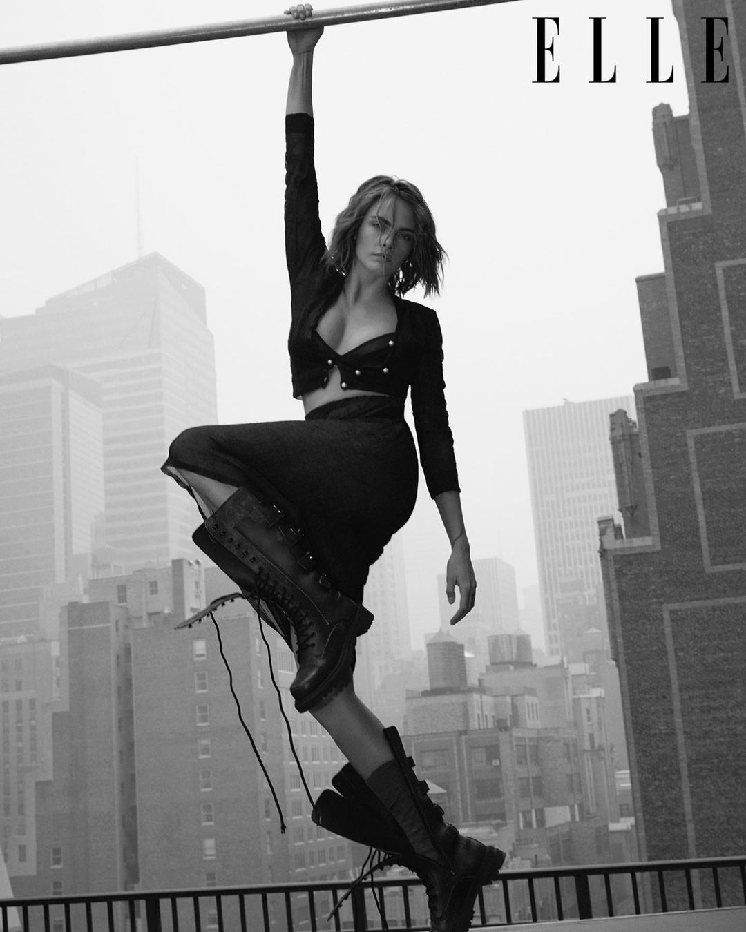 Model Cara Delevingne hanging from a high-rise rooftop bar against an urban skyline, shot in monochrome by Quentin Jones for Elle.