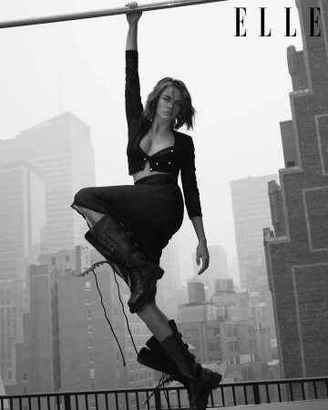 Model Cara Delevingne hanging from a high-rise rooftop bar against an urban skyline, shot in monochrome by Quentin Jones for Elle.