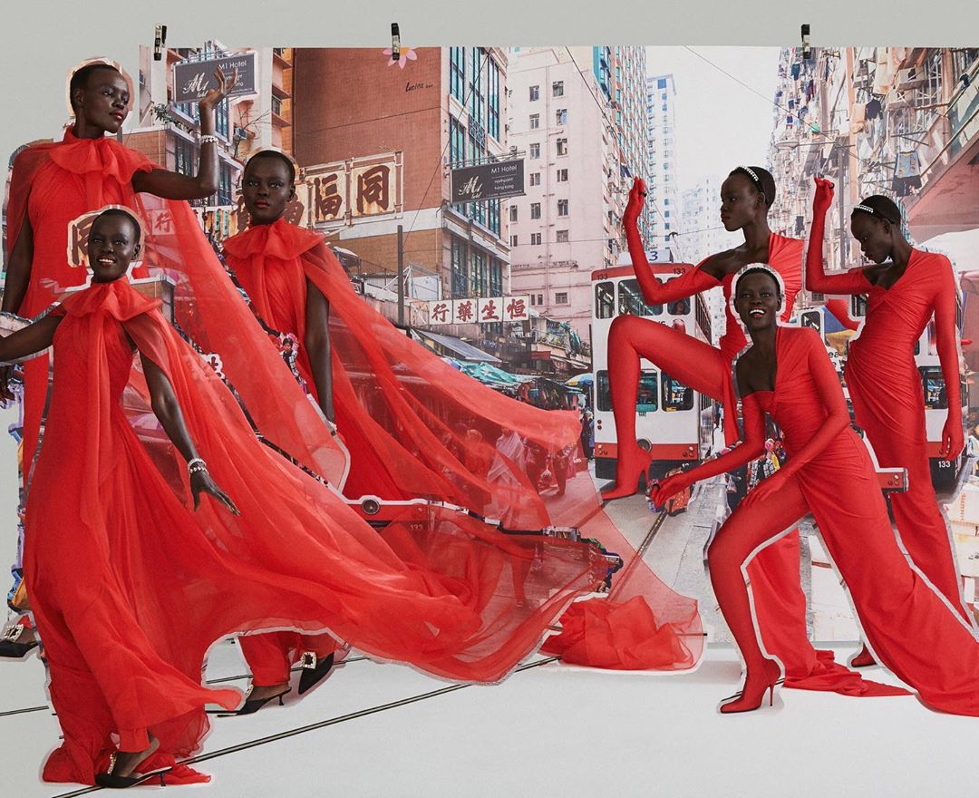 Print of multiple African models wearing red dresses and jumpsuits, posing in front of an urban street scene in daytime lighting.