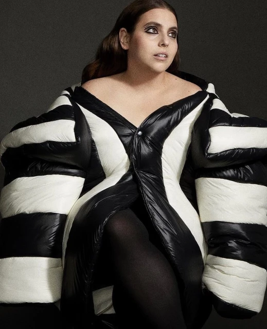 Woman poses in a studio wearing an oversized black and white striped puffer coat dress and black tights against a dark grey background.