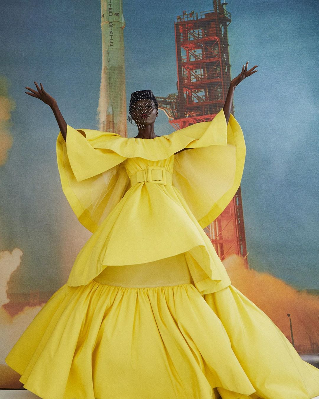 Fashion shot: A woman in a voluminous yellow gown with a veiled headpiece stands against a backdrop of a rocket launch. 