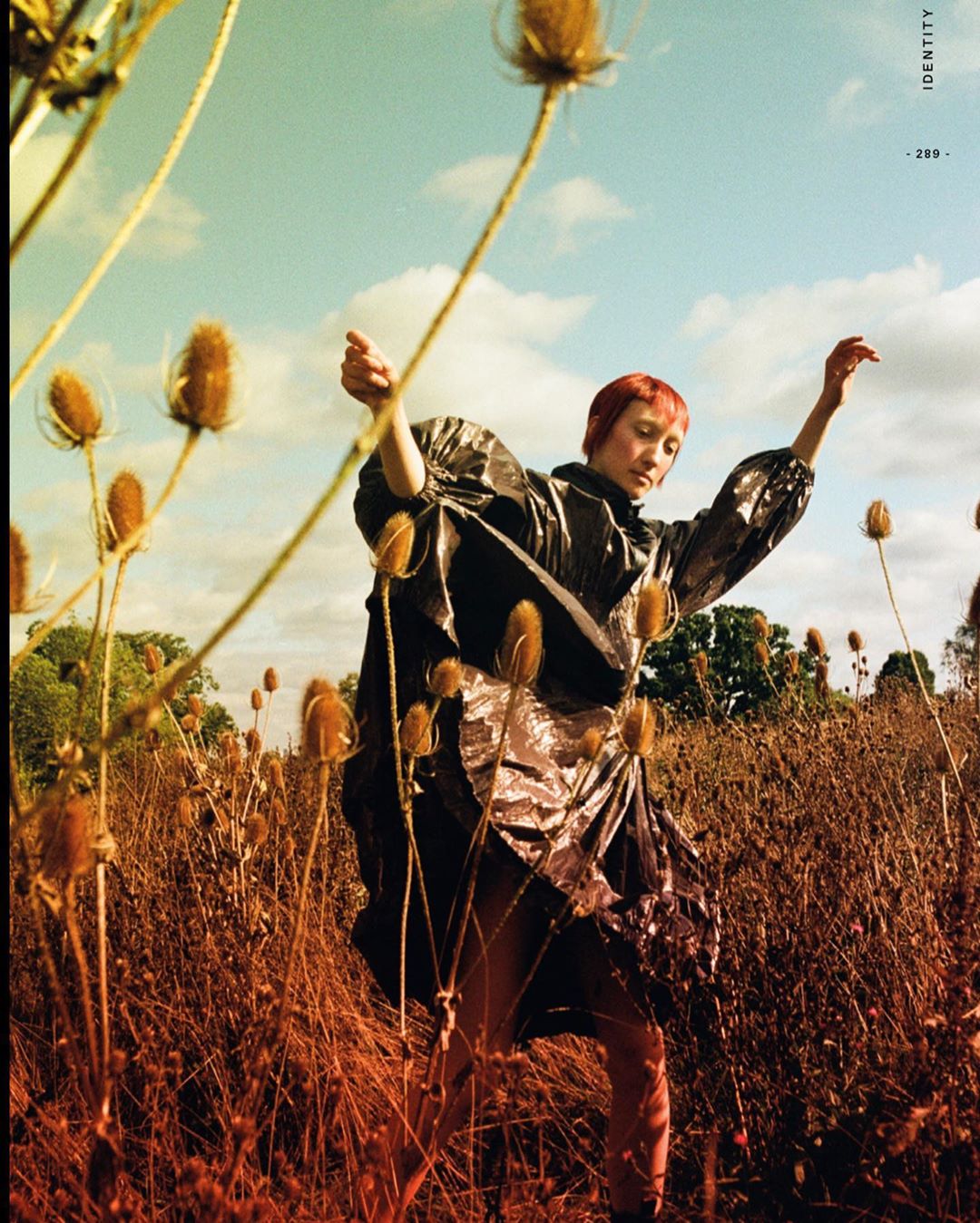 Woman in a shiny coat posing in a field of brown teasel plants under a blue sky with thin clouds, captured in an outdoor editorial shot.
