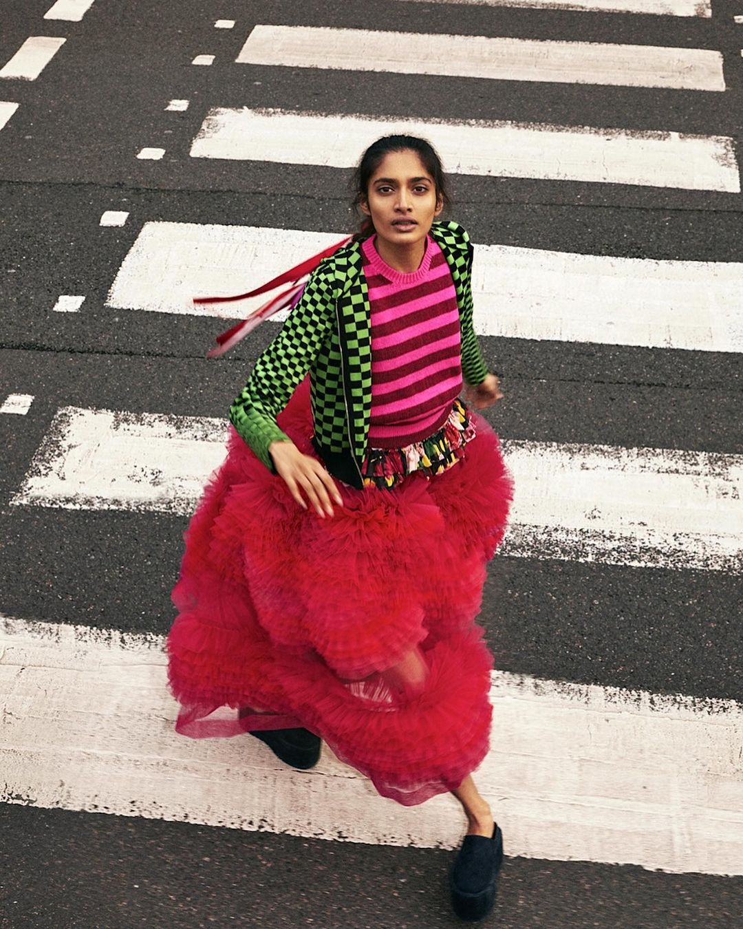 Woman in a pink tutu and checkerboard jacket is running across a crosswalk with white stripes. The view is from above.