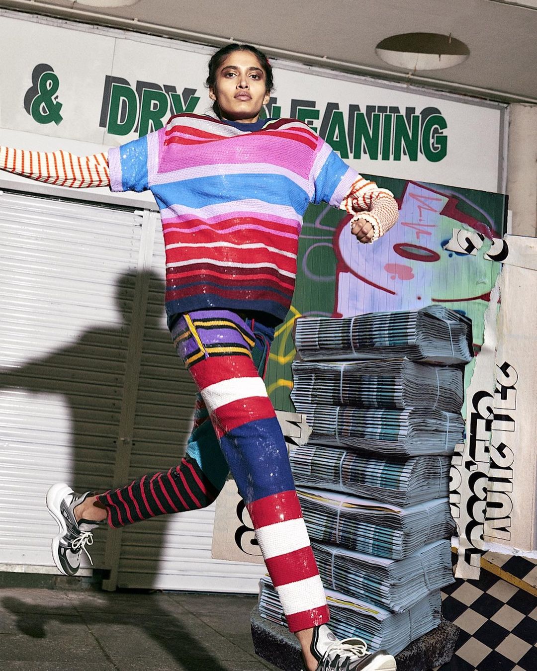 Woman in striped top and pants jumps in front of a dry cleaning business and a collage of newspapers and graffiti art.