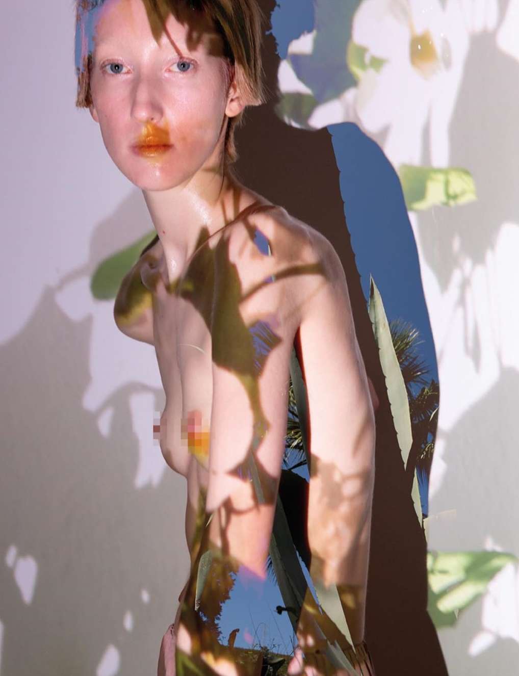 A young, fair-skinned, topless woman is captured in a studio, her body overlaid with a shadow projection of foliage and blue sky.