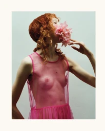 Red-haired model wearing a sheer pink mesh dress holding a pink flower to her mouth against a white background