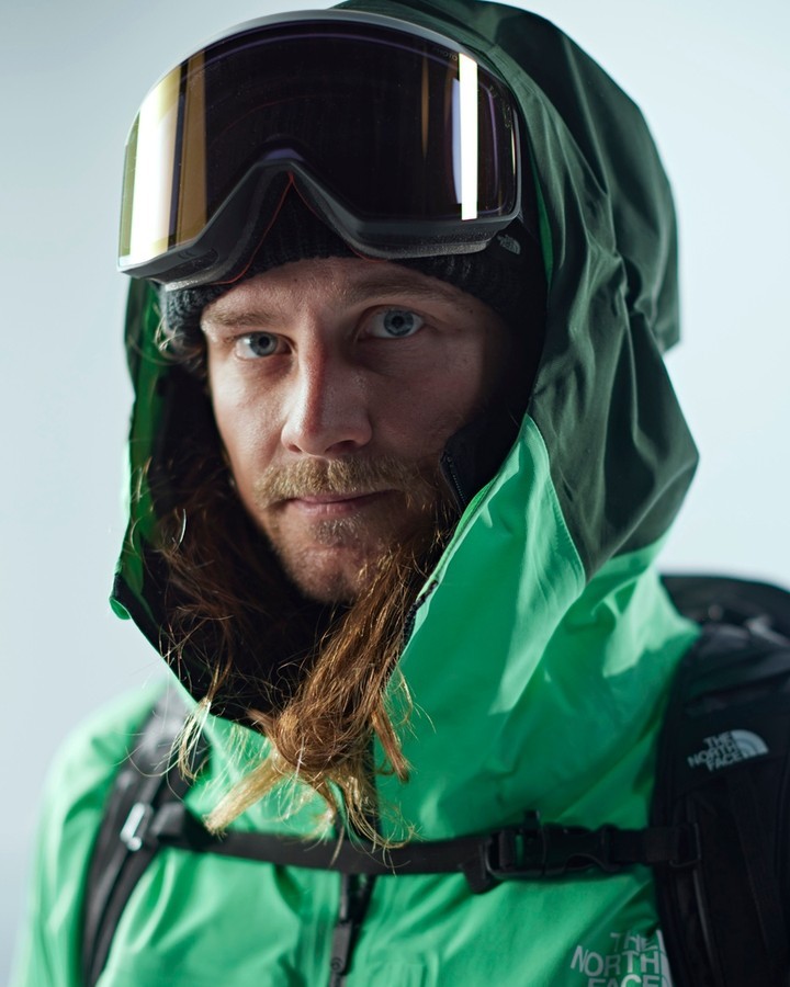 Close-up portrait of an adult man in ski goggles, wearing a green hooded jacket and black backpack, with a light background.