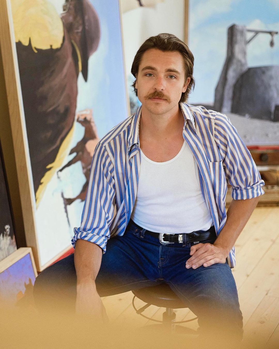 Stylish man with a mustache poses in front of paintings in a studio, wearing a striped shirt and jeans in an indoor setting.