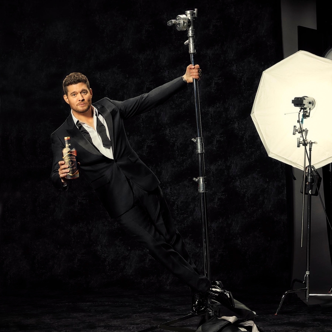 Adult male in black tuxedo holds a bottle while posing next to lighting equipment against a dark backdrop in a photography studio.
