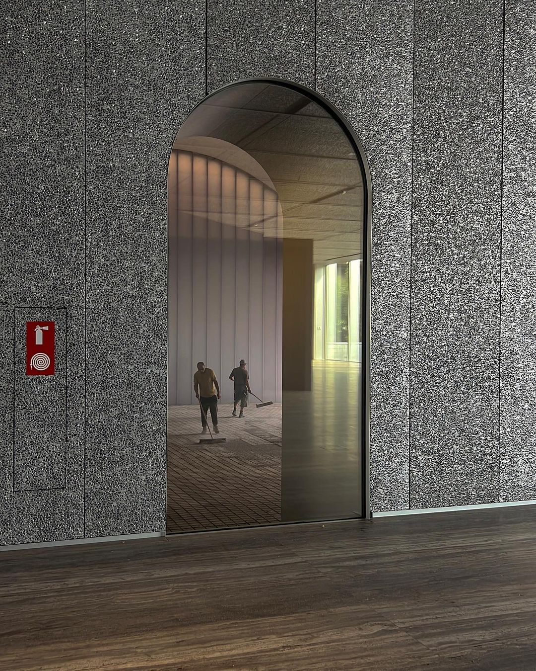 Interior shot of two maintenance workers cleaning inside a modern building, viewed through an arched doorway. Architectural textures visible.