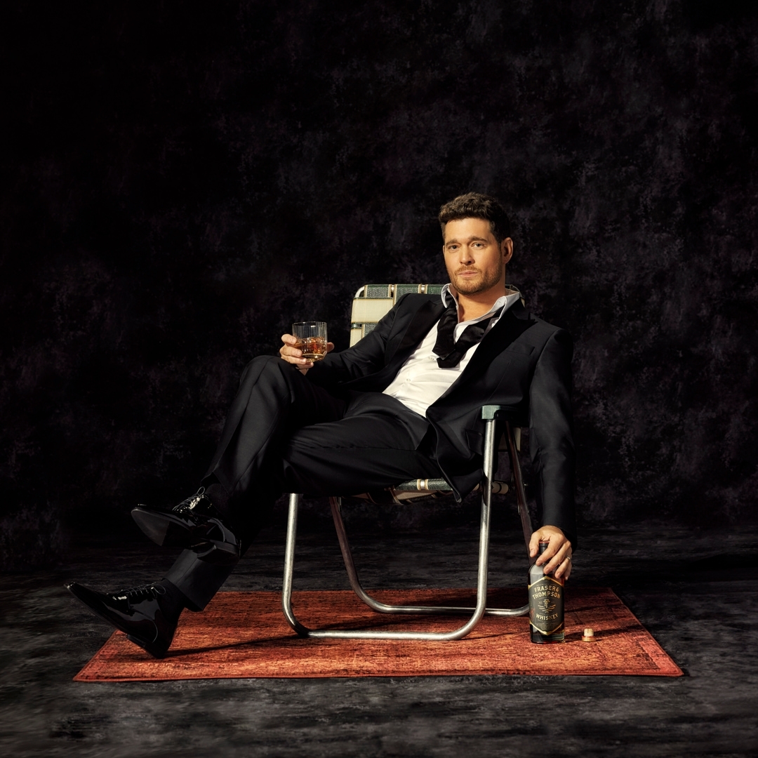 Michael Buble is seated in a folding chair wearing a suit and holding a glass of whiskey, with a bottle of alcohol on the floor.