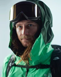 Man wearing green waterproof jacket and ski goggles with reflective visor over beanie hat