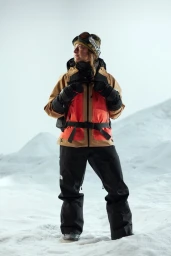 A skier in a tan, red, and black winter jacket and goggles standing on a snowy mountain landscape