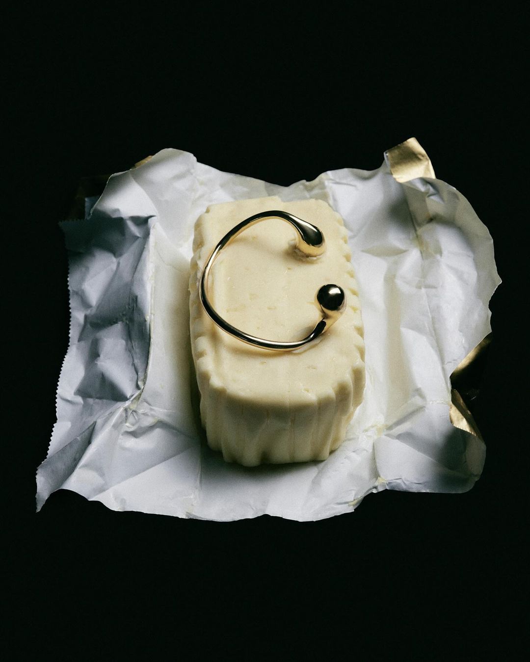 Stylized shot of a block of butter topped with a gold cuff bracelet, set against a dark background and crinkled foil wrapper.