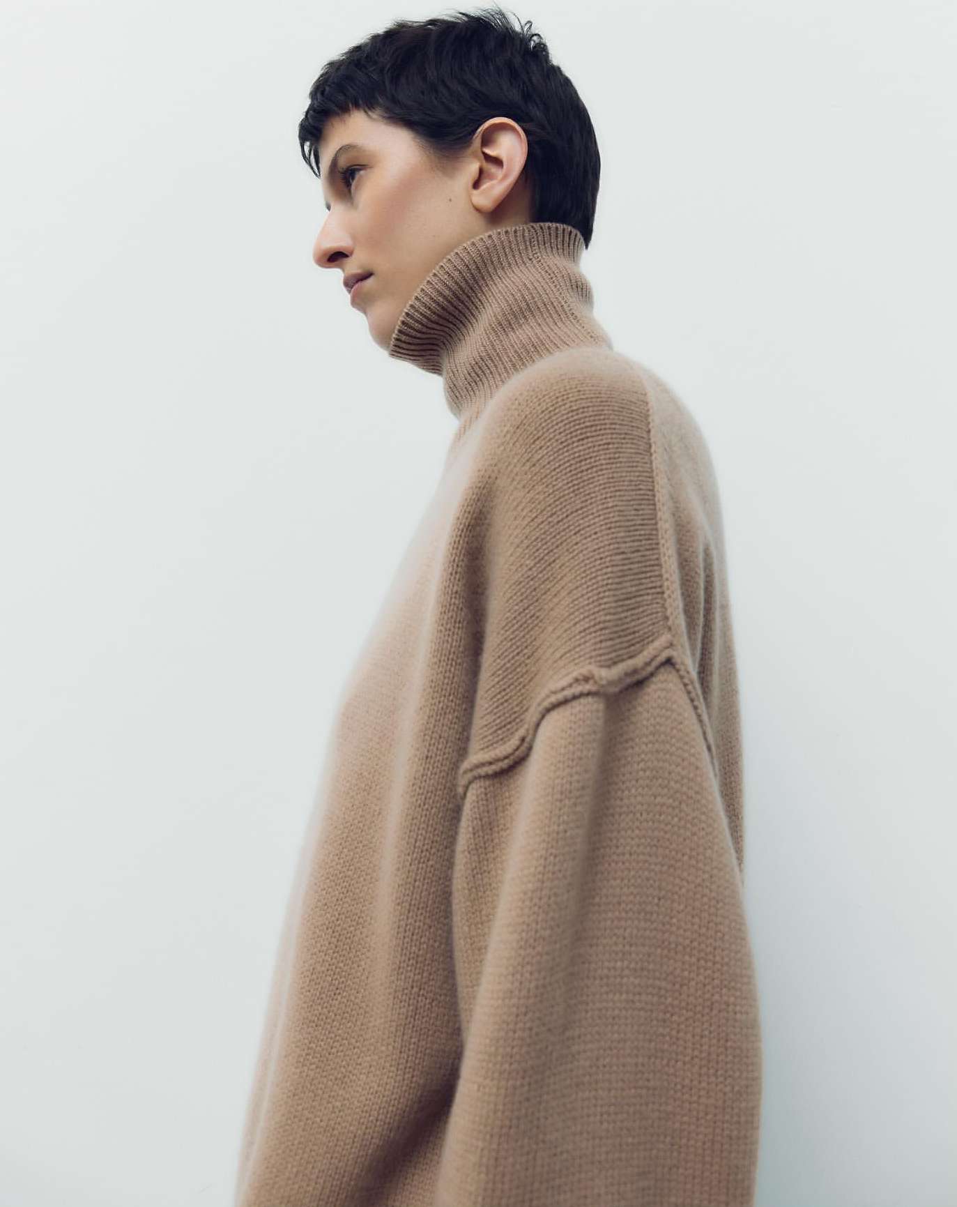 Profile shot of an adult female wearing a tan turtleneck sweater, with short black hair, against a plain white studio backdrop.