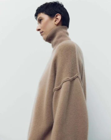 Profile shot of an adult female wearing a tan turtleneck sweater, with short black hair, against a plain white studio backdrop.
