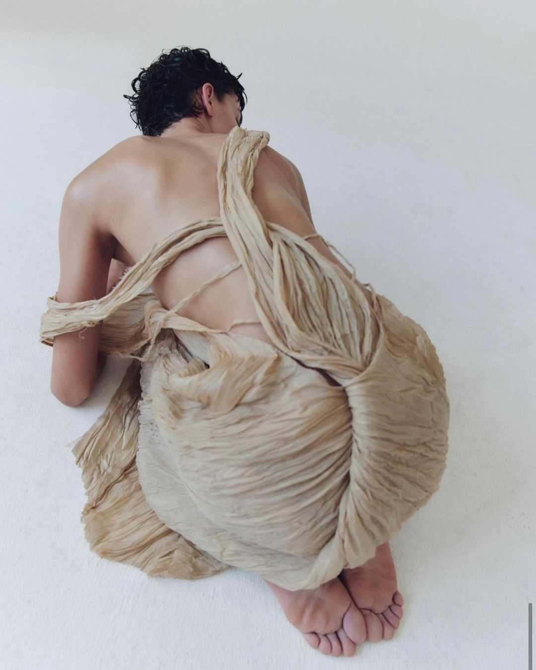 Back view of a person draped in textured beige fabric, seated on a white surface, showcasing fashion and textile design in a studio setting.