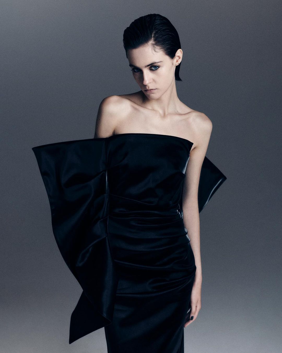 Elegant woman in a draped black gown with a large bow detail, posing against a neutral grey backdrop in a fashion studio setting.