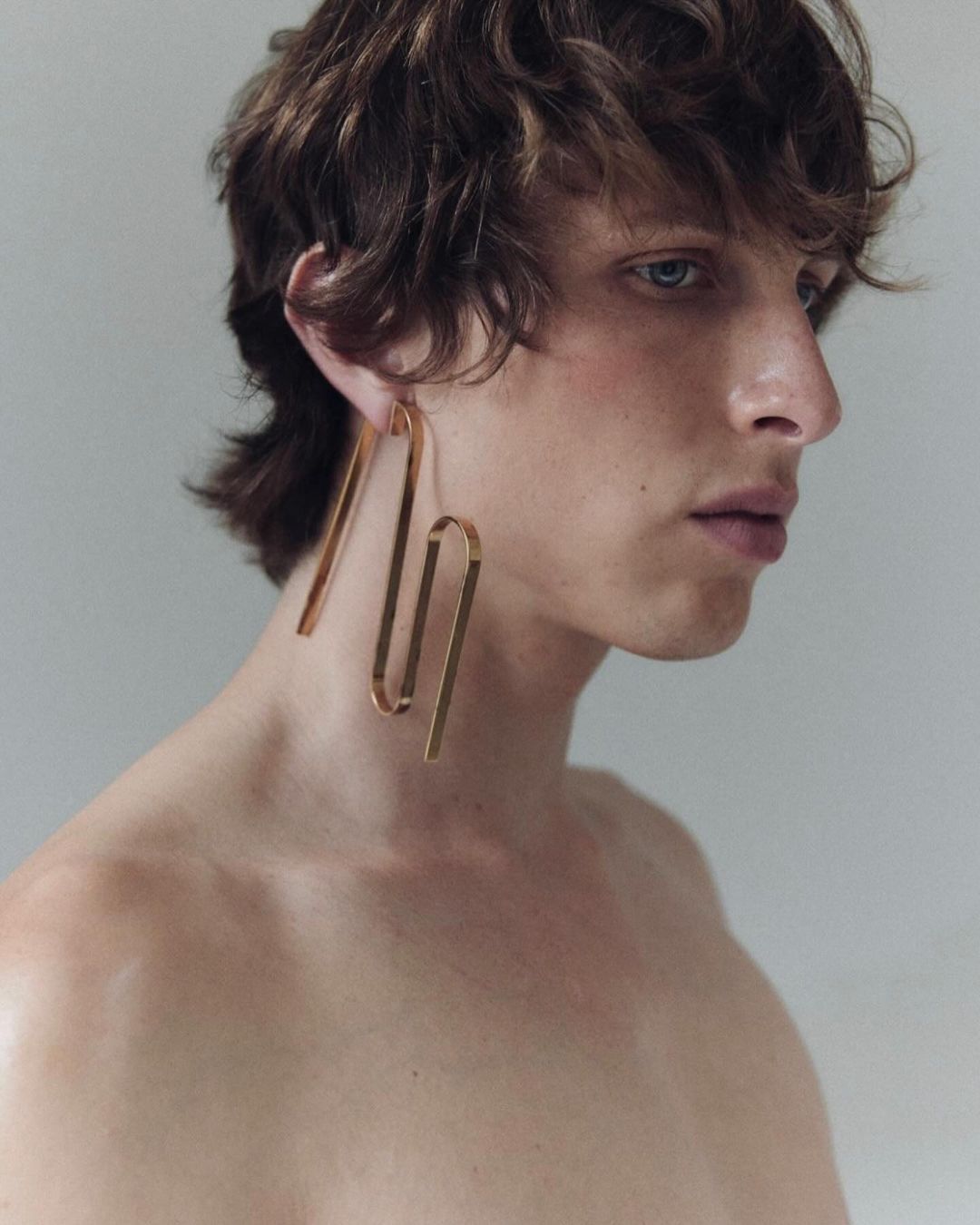 Profile portrait of a shirtless person with brown hair wearing gold geometric earrings against a neutral grey background in a photo studio.