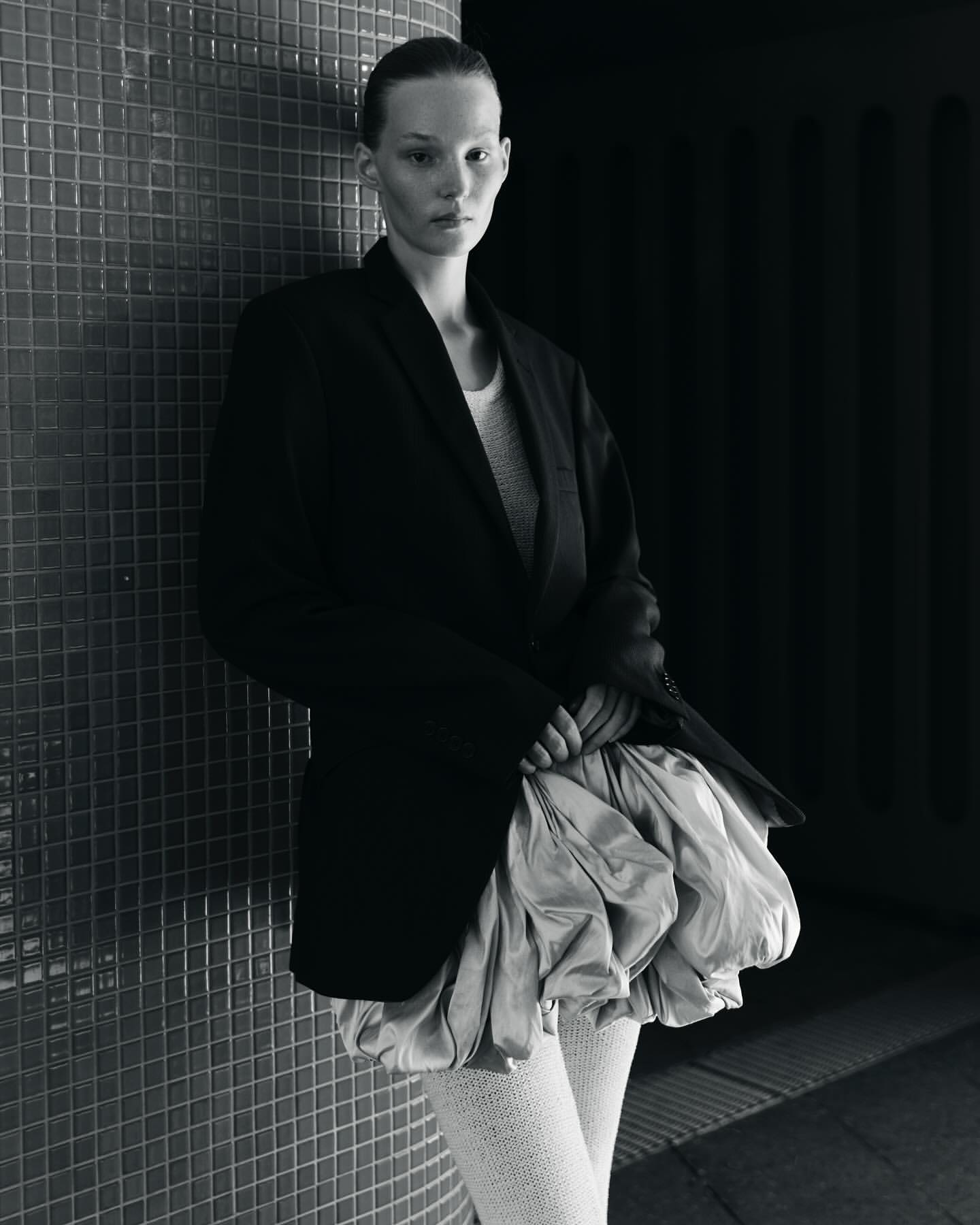 Model leaning against a dark tiled wall wearing an oversized black blazer and voluminous pale skirt, photographed in Berlin.