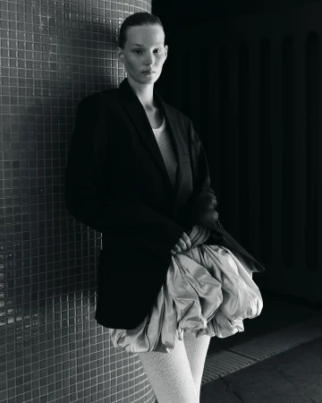 Model leaning against a dark tiled wall wearing an oversized black blazer and voluminous pale skirt, photographed in Berlin.