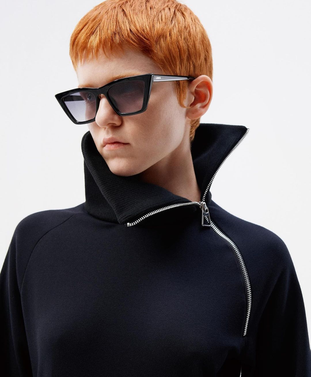 Portrait of a woman with red hair and black sunglasses wearing a navy blue sweatshirt with a high collar and a silver zipper.