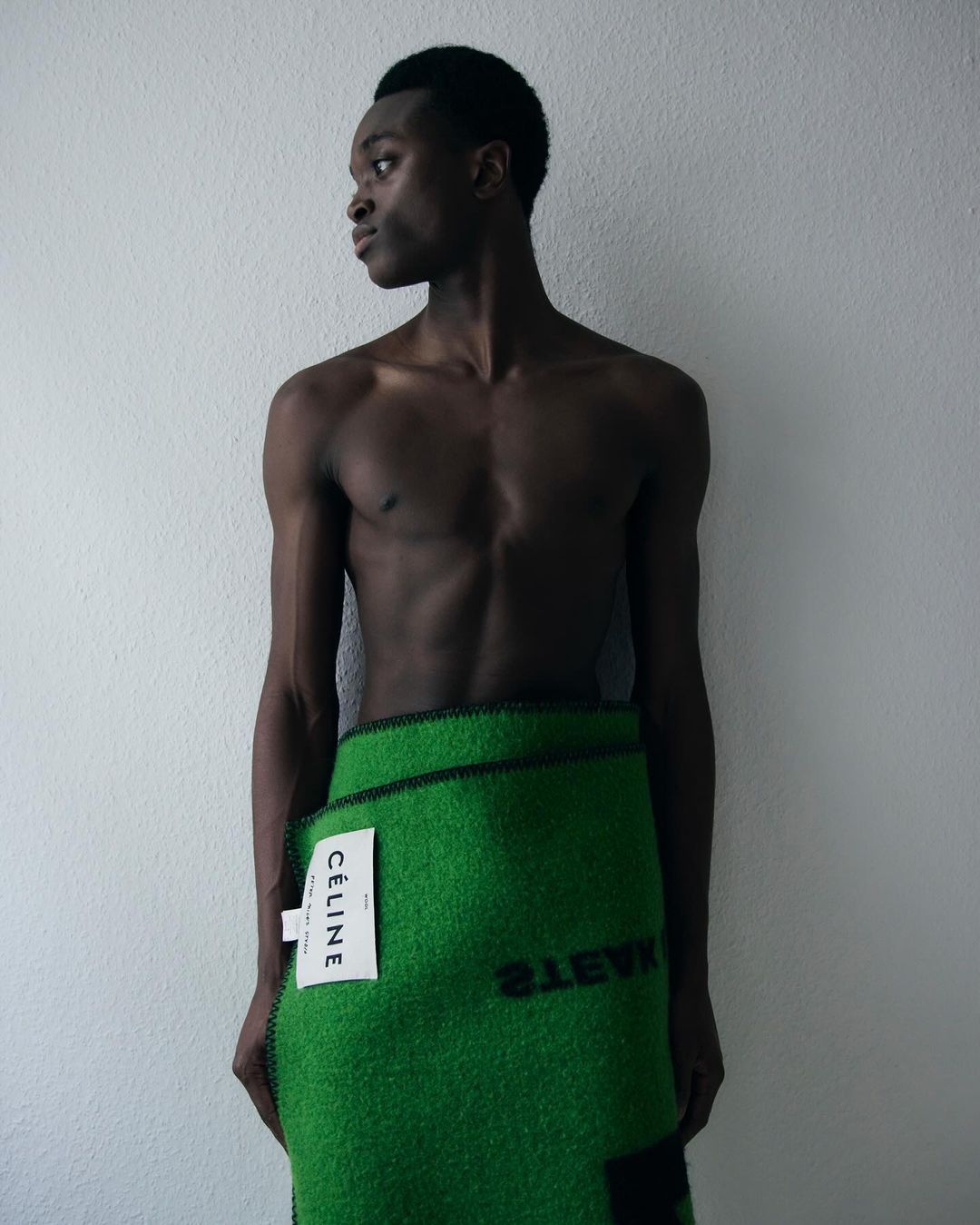 Adult male poses indoors wearing a green textile material with 'WASTE' text and 'CELINE' logo. Studio lit portrait against plain background.