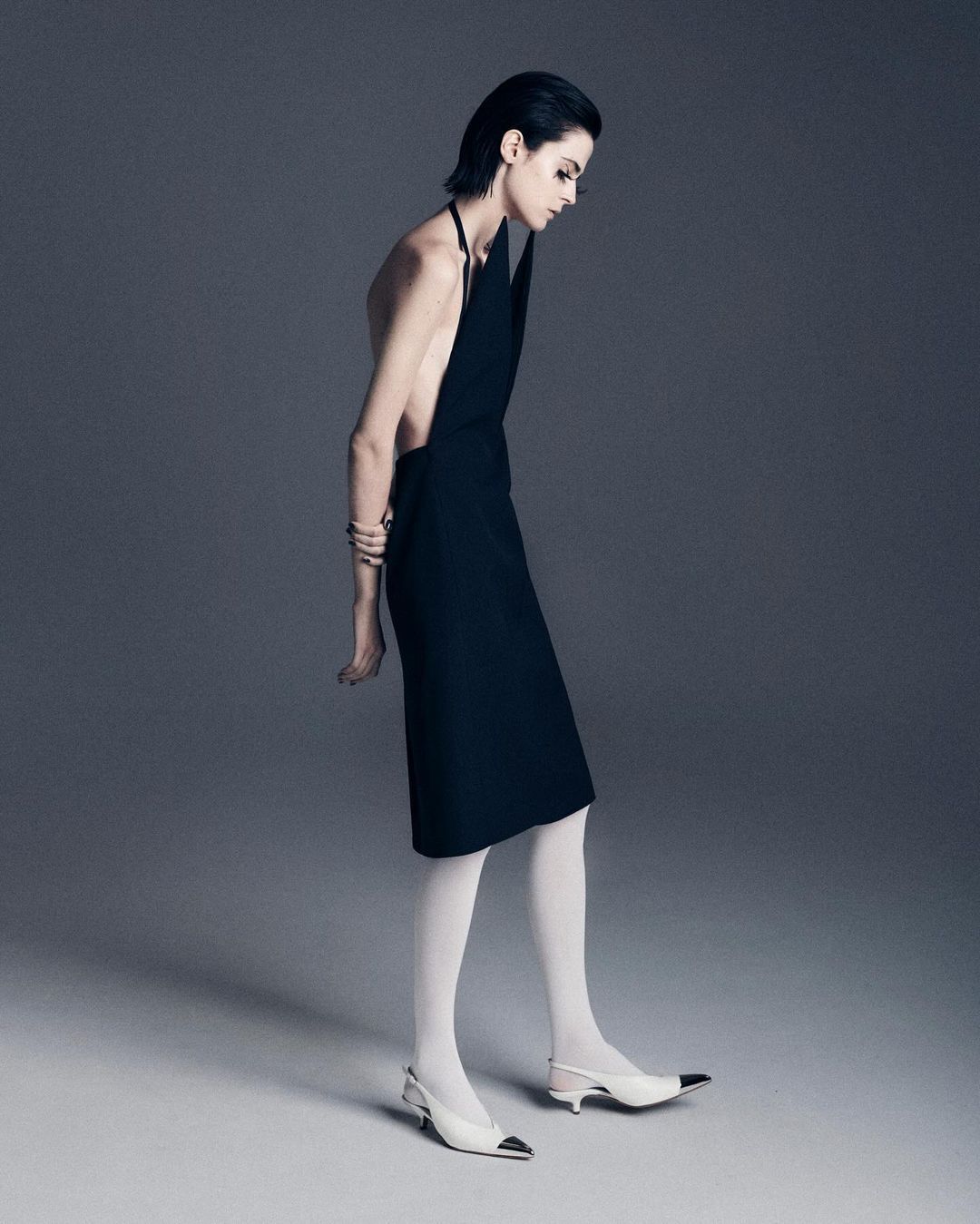 Woman in black halter dress and white heels poses in a studio with a neutral gray background in a fashion studio shot.
