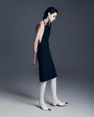 Woman in black halter dress and white heels poses in a studio with a neutral gray background in a fashion studio shot.