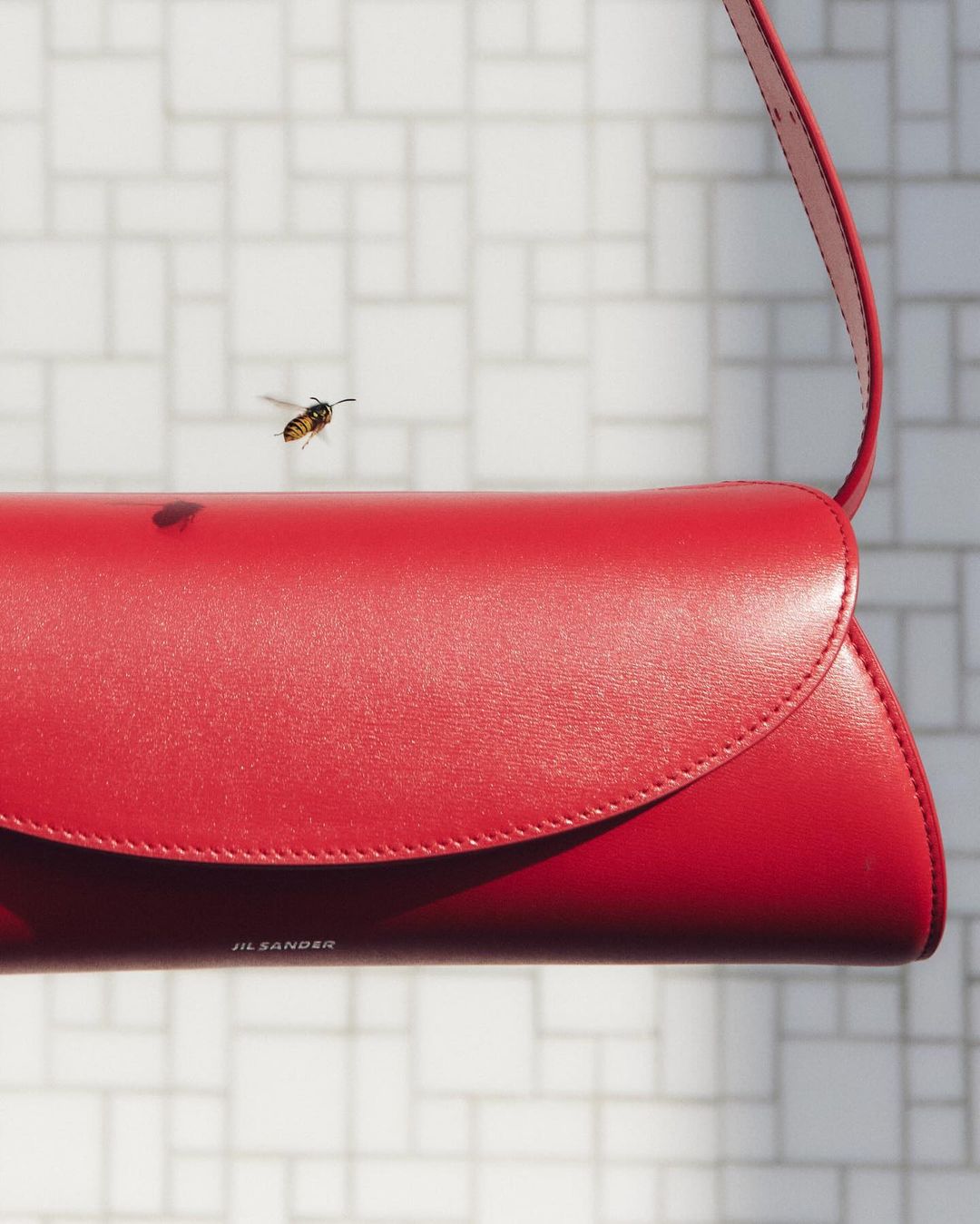 Red leather Jil Sander handbag floats with a bee in a studio setting against a modern, white-tiled background, product shot.