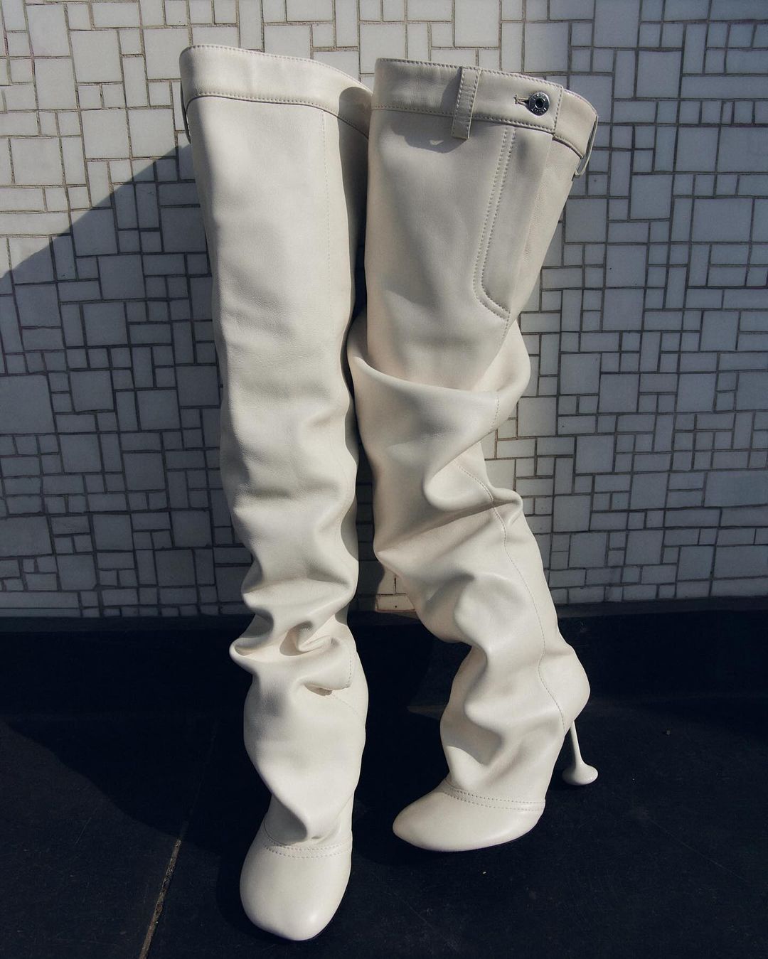 Two white leather thigh-high heeled boots are standing, one slightly slumped, in a studio setting against a mosaic tile background.