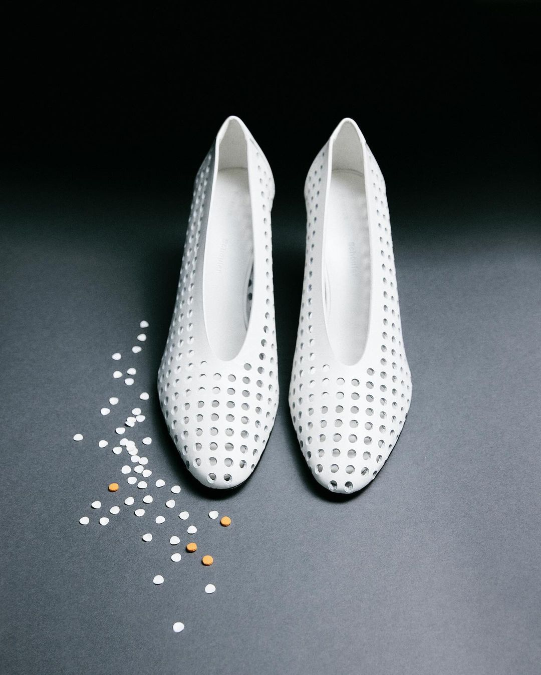 Pair of white, perforated high heels on a dark gray surface, scattered with small white and tan round objects, creating a minimalist fashion product shot.