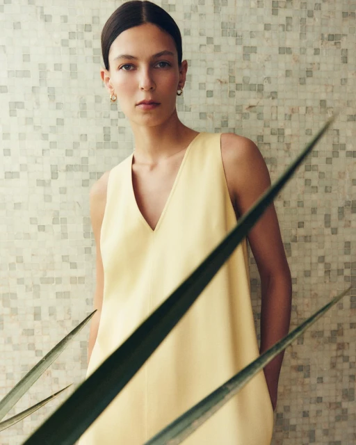 An adult female stands in front of a tiled wall, wearing a yellow sleeveless dress, with green plant leaves in the foreground.