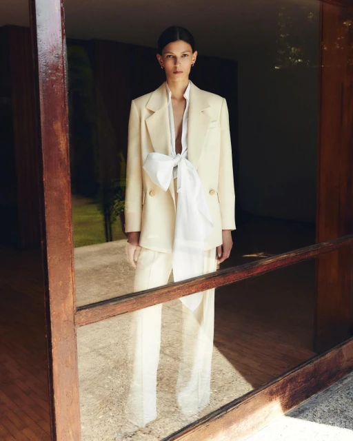 Fashionable woman in an ivory blazer and matching pants, posed in front of an open door with a blurred, outdoor background.