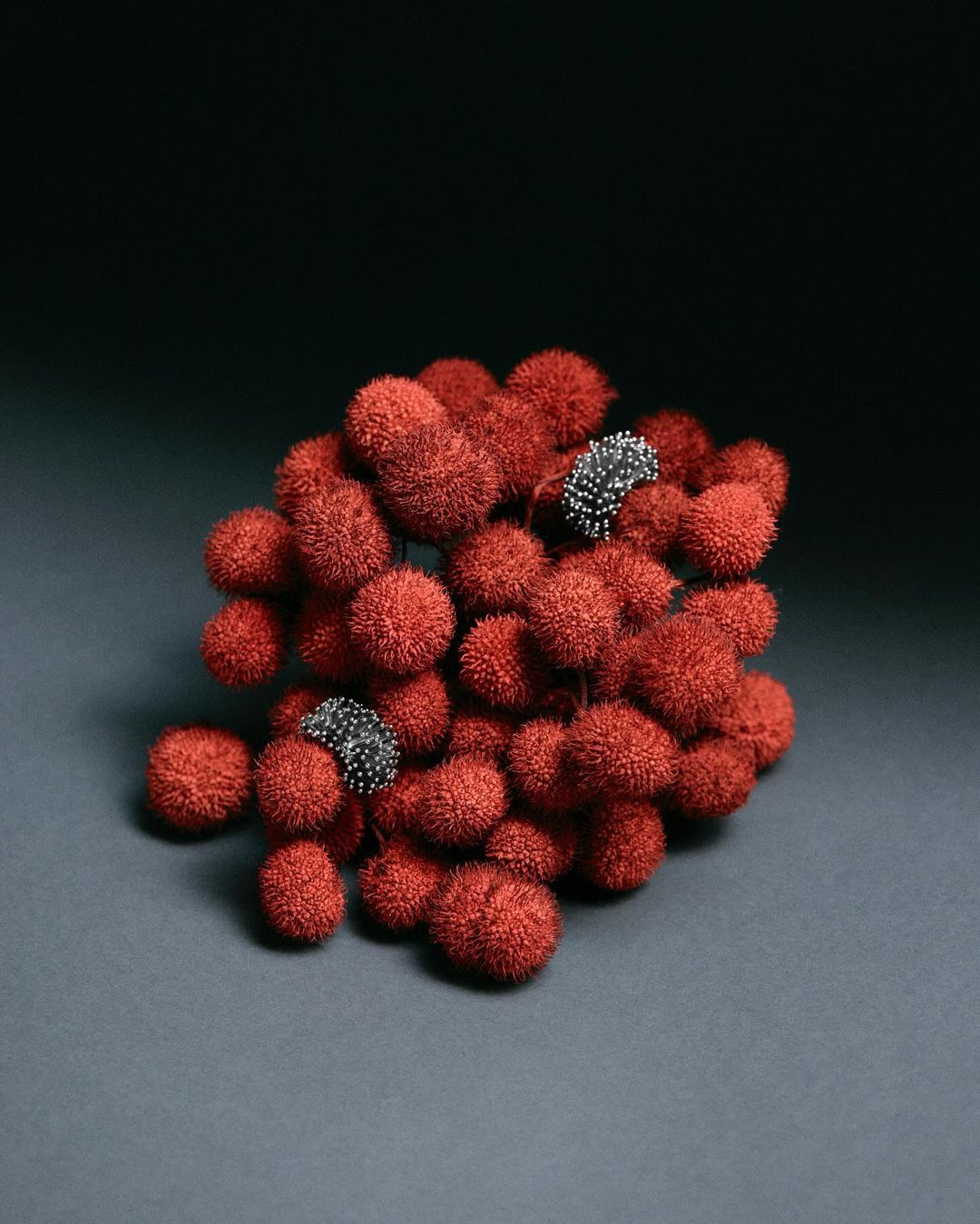 Close-up studio shot of an abstract red cluster of spiky round forms, with two smaller clusters of gray/silver spikes on a grey background.