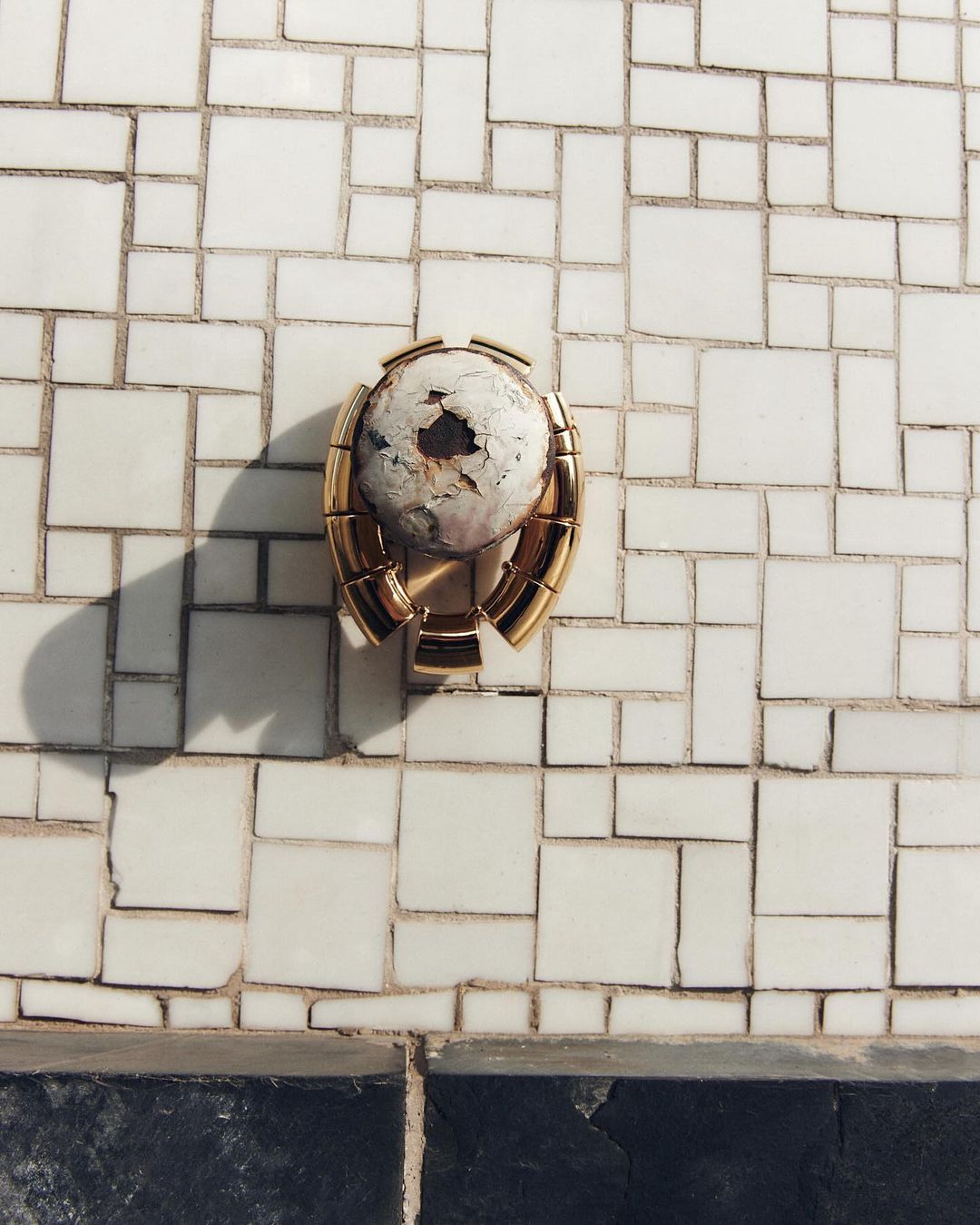A wall with a mosaic tile pattern features a metallic gold bracket holding a distressed white sphere, creating an aged, textured look.