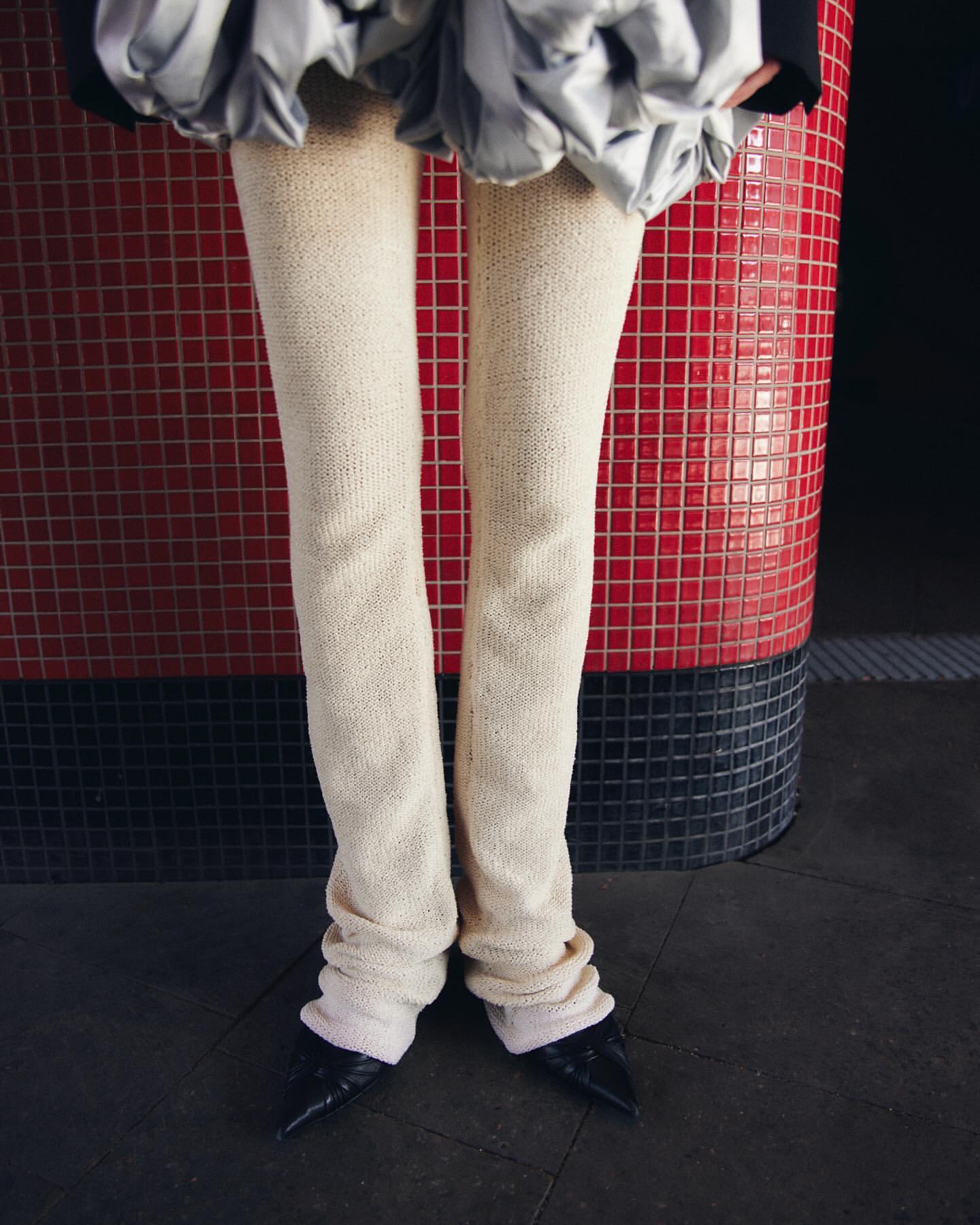 Fashionable leg warmers and pointed-toe heels worn indoors, standing against a red-tiled background for a chic, contemporary aesthetic.