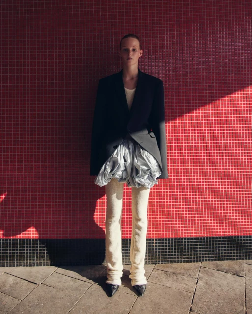 Fashionable woman poses in a blazer, silver skirt, and cream-colored leggings against a red tiled wall with shadows and natural lighting.