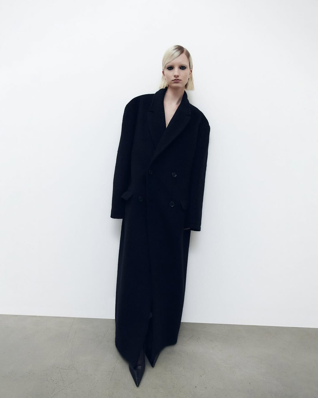 Fashion shot of a woman in a long black coat against a white background, showcasing a minimalist aesthetic and bold silhouette.