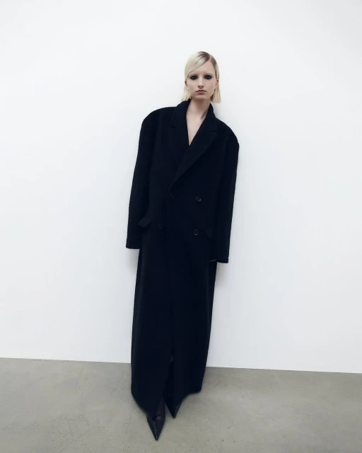 Fashion shot of a woman in a long black coat against a white background, showcasing a minimalist aesthetic and bold silhouette.